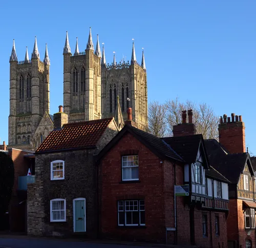 In this urban architectural photograph, Lincoln Cathedral rises prominently above a row of traditional houses in Lincoln, England. The iconic towers and spires of Lincoln Cathedral, a famous landmark in the United Kingdom, are illuminated by the clear midday winter sunlight, casting sharp lines against the blue sky. The setting, with its mix of historic red brick and stone houses in the foreground, highlights the rich architectural heritage of Lincoln, while the cathedral dominates the skyline as a testament to the city’s medieval past. The image captures the crisp, bright atmosphere characteristic of a winter afternoon in England, making Lincoln Cathedral the unmistakable focal point of this picturesque scene.
